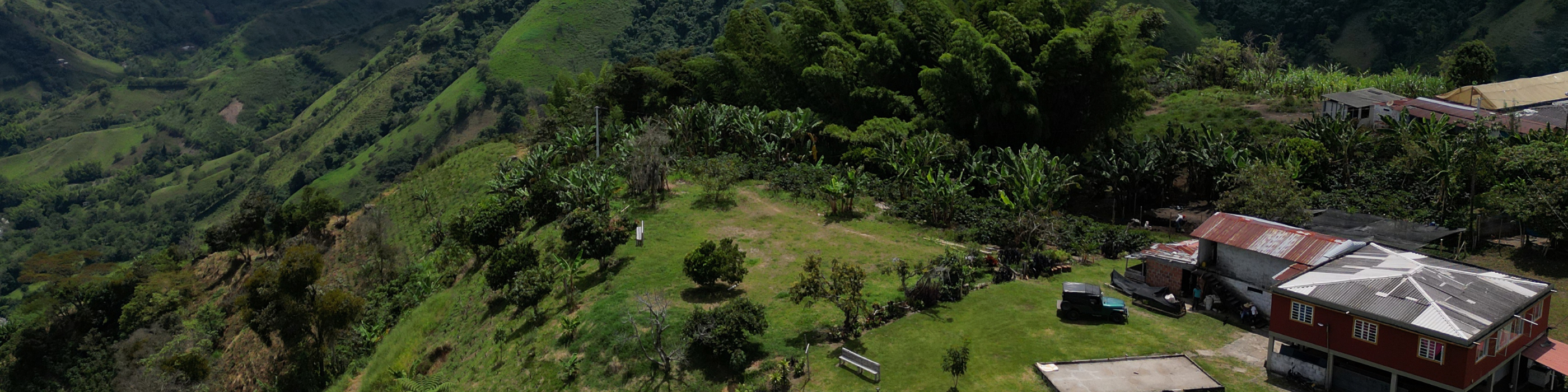 Blick aus der Luft auf die Finca El Binomio, gelegen an einem Hang inmitten von Kaffeeplantagen, mit einem roten Haus, rustikalen Gebäuden und tropischer Vegetation.